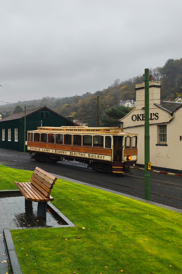 Photographie, sous un ciel sombre, d’un train électrique portant la mention <q>Douglas Laxey & Ramsey Electric Railway</q>. Le train est jaune et marron, d’un style très ancien, et composé d’un seul wagon. Au devant, un banc, et à l’arrière, quelques maison puis les montagnes.