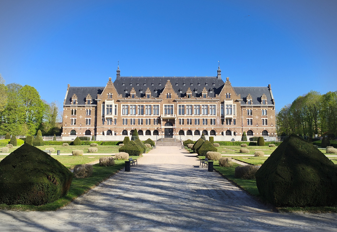 Photographie d’un château imposant, fait de briques rouges et avec un toit noir. Au premier plan, un parc bien entretenu avec des arbres taillés au carré.