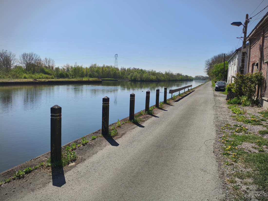 Photographie d’une route, au premier plan. Au second plan, le long de la route, un cours d’eau calme. À l’arrière-plan, on aperçoit l’autre rive, avec de nombreux arbres. Sur le bord de la route, une maison et une voiture garée