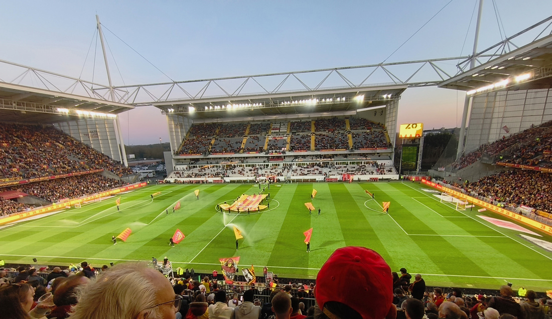 Photographie de l’intérieur d’un stade de football. Au premier et à l’arrière-plan, on voit une masse de supporters. Au centre, le terrain sur lequel de nombreuses personnes ont des drapeaux et des maillots rouges et jaunes.