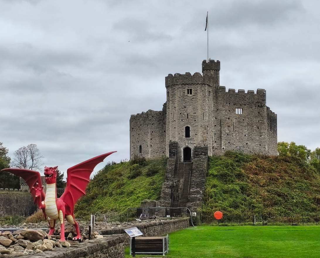 Photographie d’un monument de pierres grises, avec des remparts semblables à ceux d’un château-fort, quelques fenêtres creusées comme celles de prisons, et un petit escalier de pierre pour y accèder. Au devant triomphe un dragon rouge avec la patte droite levée.