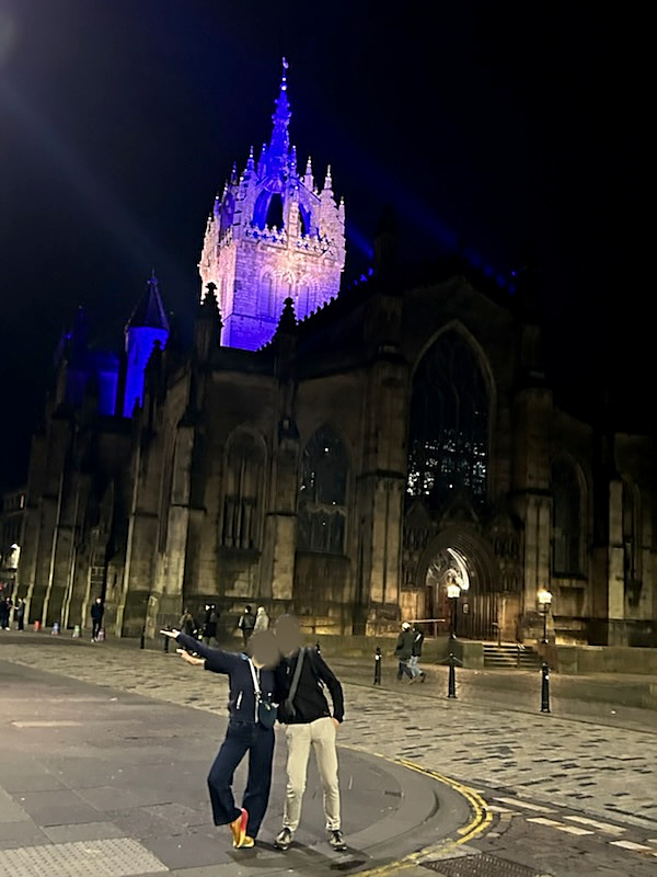 Photographie de nuit d’une cathédrale que l’on distingue mal dans son ensemble, mais dont la pointe est éclairée en bleu. Au premier plan, deux personnes posent.